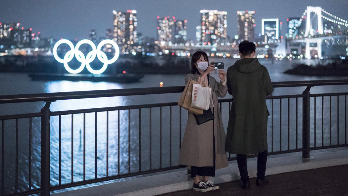 A Couple taking a selfie at night in front of a neon-lit olympic logo sign.