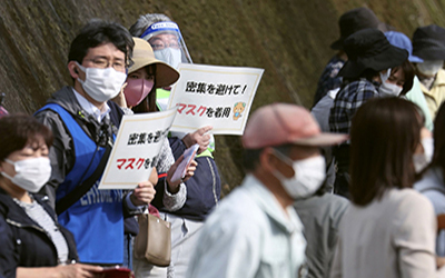 Some audience members carry signs in japanese while wearing masks. Translated, the signs say 'Avoid Crowding, Wear Masks!'