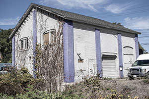 A white and periwinkle house on the side of an intersection with plantlife growing over it.